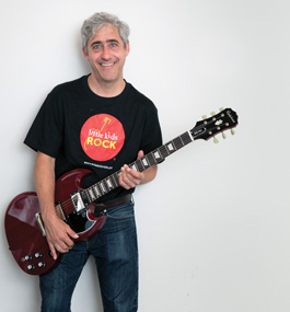 A man wearing a Little Kids Rock t-shirt holds an electric guitar